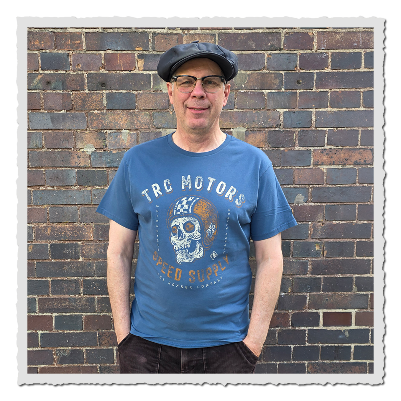 Frontansicht eines Mannes mit blauem Grafikshirt, dunkler Hose und Kappe vor rustikaler Ziegelwand.